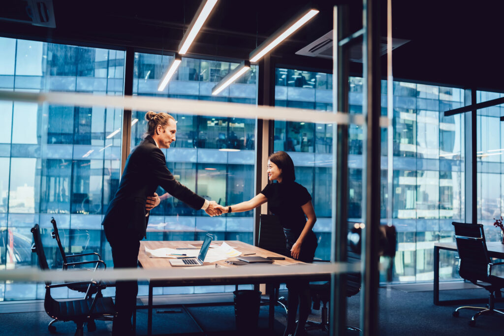 Image of a male and a female business professional shaking hands across a table in an office building.