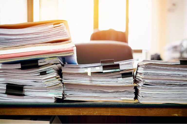 Large stacks of paperwork, folders, and documents piled high on a desk in front of an empty office chair, symbolizing work backlog and the operational cost of an unfilled job position.