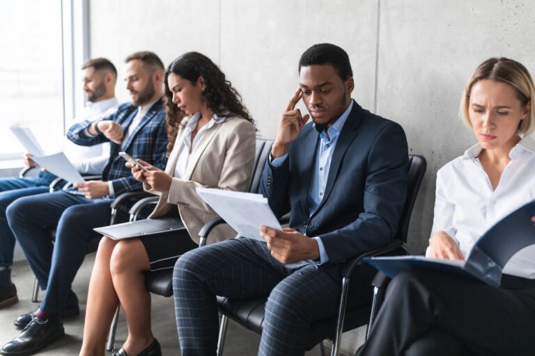 A diverse group of five professionals sitting in a row in a waiting area, anxiously reviewing resumes and checking their devices before a job interview.