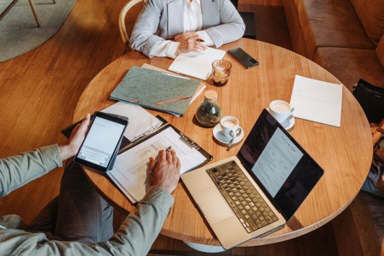 Top-down view of hiring managers and recruiters collaborating at a round wooden table with laptops, tablets, and candidate resumes.